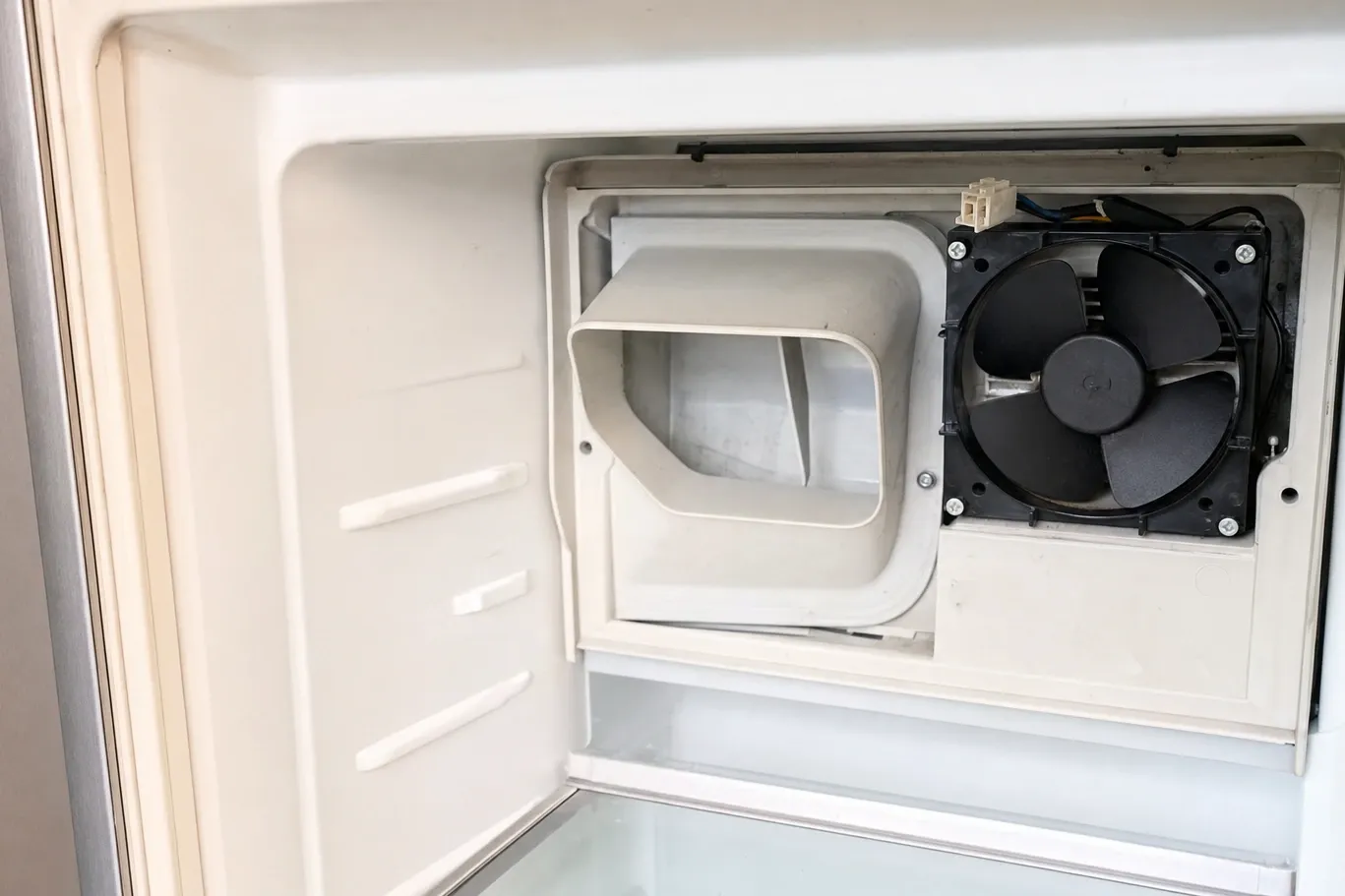The rear duct cover removed inside an empty freezer compartment, exposing the evaporator fan in its housing beside the open air duct