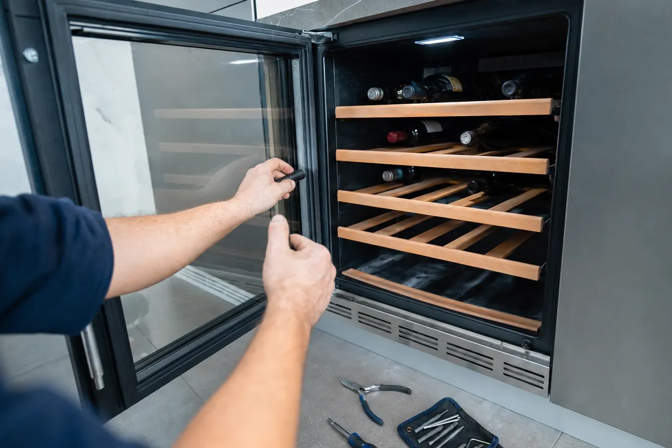An undercounter wine cabinet with its glass door swung wide onto wooden racks, hands working at the open door edge and a pouch of nut drivers unrolled on the floor